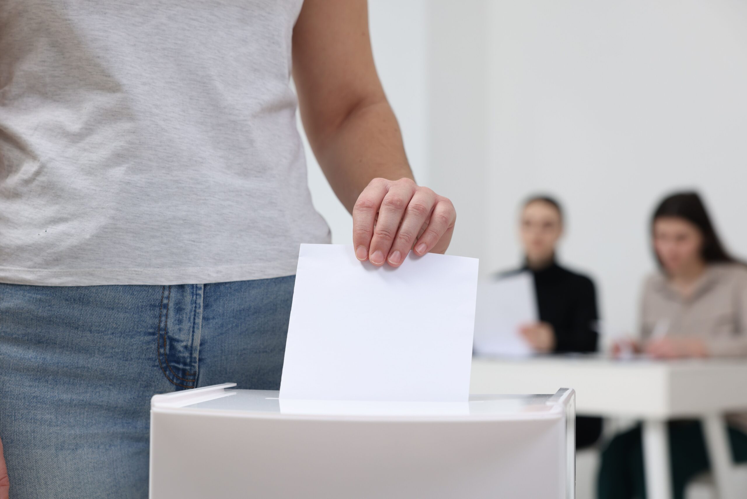Woman putting her vote into ballot box on blurred background, closeup