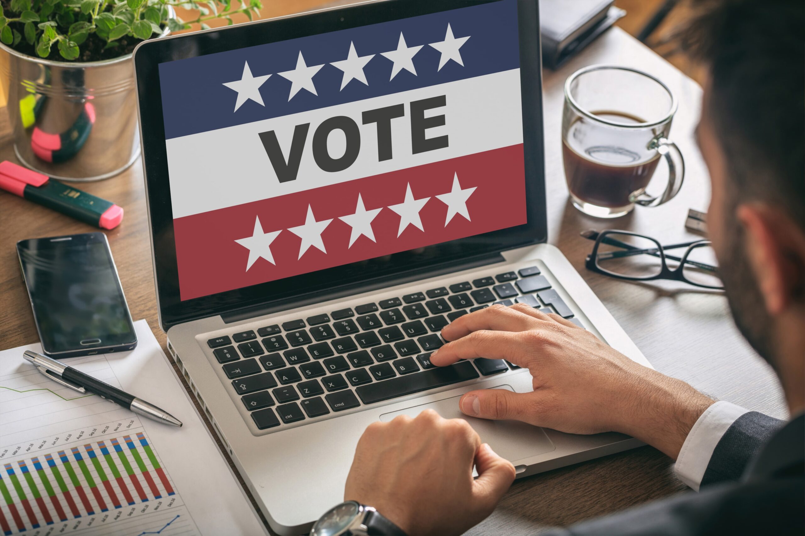A campaign staffer uses political campaign tools on a computer with a “VOTE” screen in a busy office.